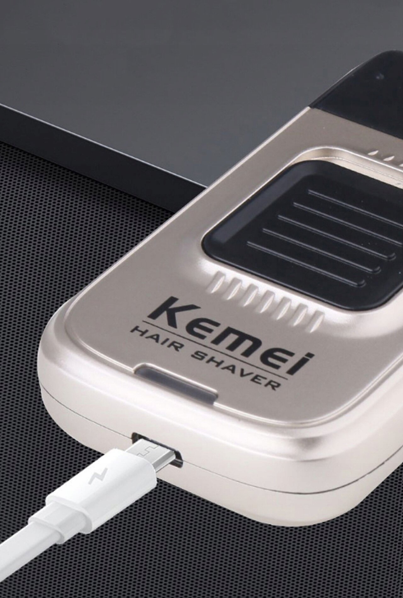 Na obrazie widoczna jest elektryczna golarka foliowa z napisem \'Kemei Hair Shaver\', podłączona do kabla USB-C. Golarka ma metaliczny kolor i prostokątny kształt.