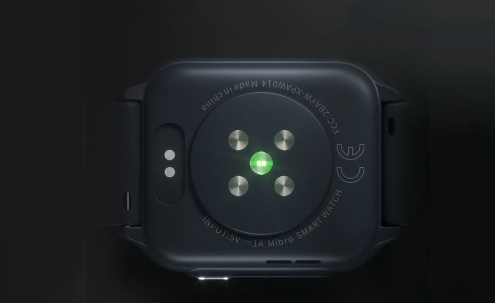 Widok tylnej części czarnego smartwatcha z czterema metalowymi sensorami i zielonym światłem. Na obudowie widoczny jest napis: \'FCC ID: 2AWX7-XPAW004 Made in China VI-1 Mibro SMART WATCH\'.