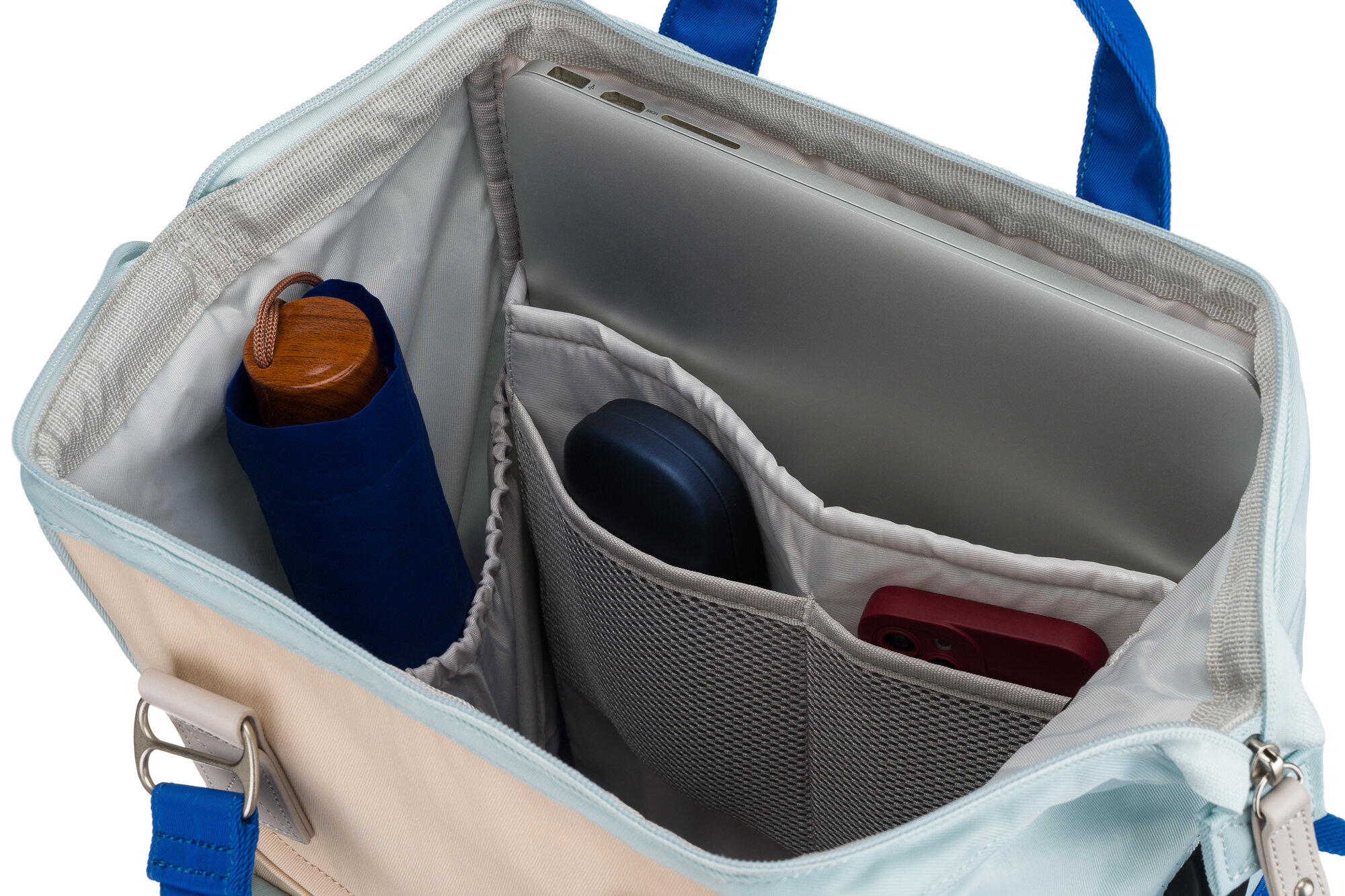 Wnętrze plecaka z widocznymi przegródkami, w których znajdują się laptop, niebieski parasol, mysz komputerowa i czerwony przedmiot. Plecak ma kremowo-niebieskie wykończenie i niebieskie uchwyty.
