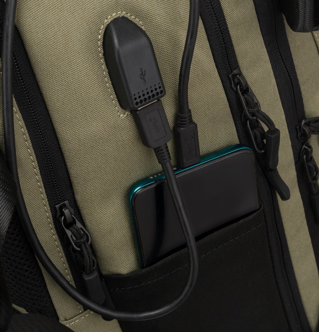 Zielony plecak z czarnymi zamkami błyskawicznymi i kieszenią, w której znajduje się telefon. Plecak posiada zewnętrzne złącze USB z podłączonym kablem.
