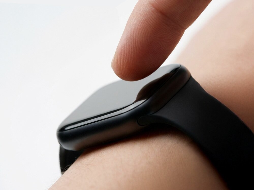 Na zdjęciu widać czarny smartwatch na nadgarstku, zbliżający się do niego palec. Urządzenie ma prostokątny ekran i czarny pasek.
