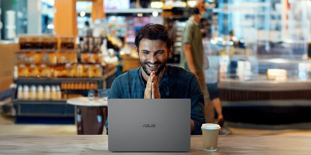 Osoba siedzi przy stole z laptopem marki ASUS, obok stoi kubek z napojem. W tle widać wnętrze sklepu z półkami pełnymi produktów.