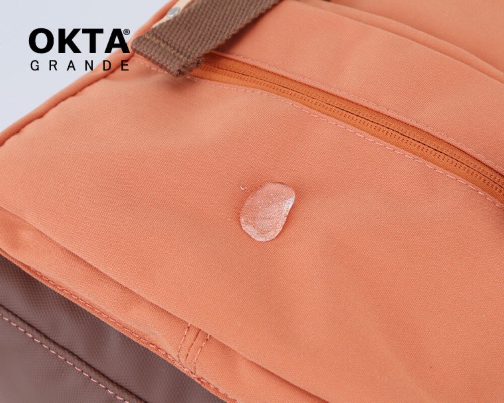 Zbliżenie na pomarańczowy materiał plecaka z widoczną kroplą wody oraz brązowym paskiem. W lewym górnym rogu znajduje się logo \'OKTA GRANDE\'.