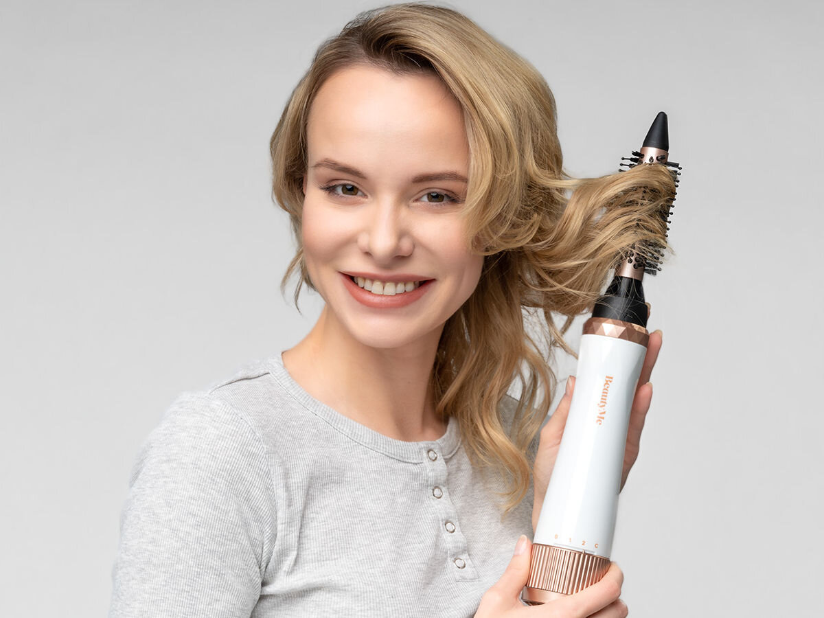 Osoba trzyma suszarko-lokówkę z widocznym napisem \'beautyme\', używając jej do stylizacji włosów. Urządzenie ma biały korpus z elementami w kolorze miedzianym.