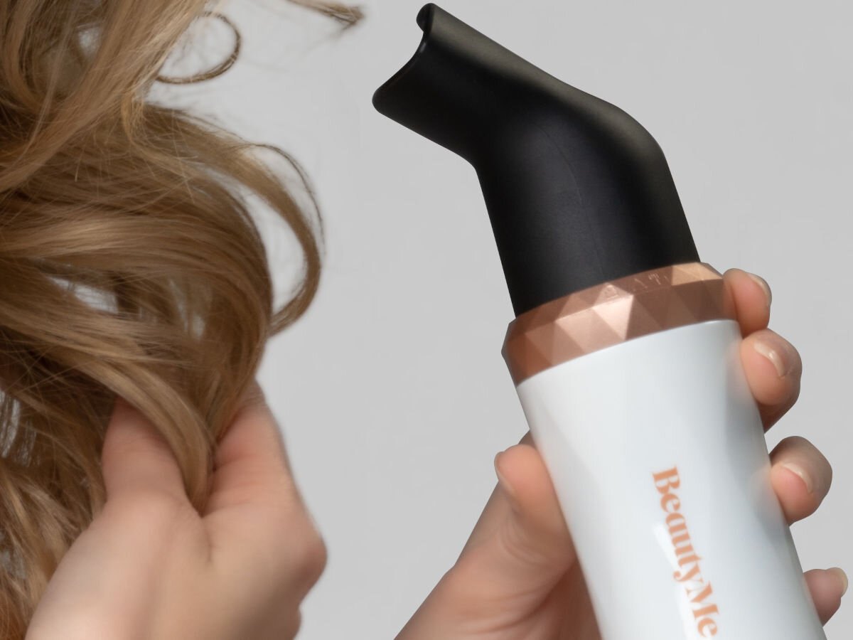Osoba trzyma suszarko-lokówkę z napisem \'BeautyMe\', z widoczną końcówką w kolorze czarnym i złotym. Obok widoczne są falowane włosy.