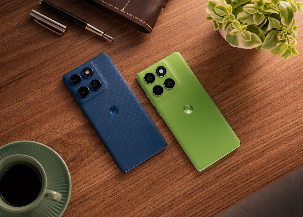 Na drewnianym stole leżą dwa smartfony, jeden w kolorze niebieskim, drugi zielonym, oba z trzema aparatami z tyłu. Obok znajduje się filiżanka z kawą, długopis, skórzane etui i roślina w doniczce.
