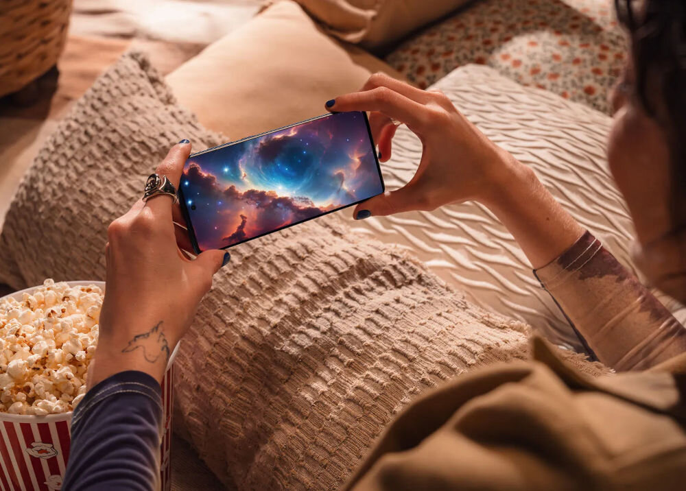 Osoba trzyma smartfon, na którym wyświetlany jest obraz kosmosu z kolorowymi chmurami i gwiazdami. W tle widoczne są poduszki i pudełko z popcornem.