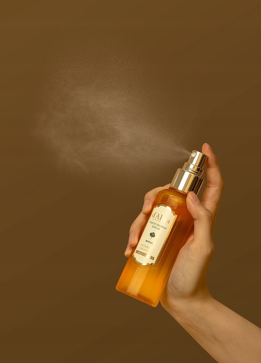 Ręka trzyma butelkę z pompką, z której wydobywa się mgiełka. Na etykiecie widoczny jest napis \'d\'Alba White Truffle Spray Serum\'.