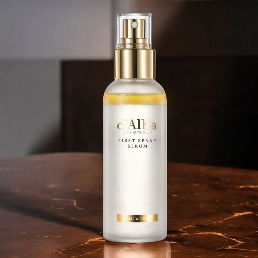 Na obrazie widoczna jest butelka z napisem \'d\'Alba piedmont FIRST SPRAY SERUM\'. Butelka ma złoty dozownik i stoi na marmurowej powierzchni.