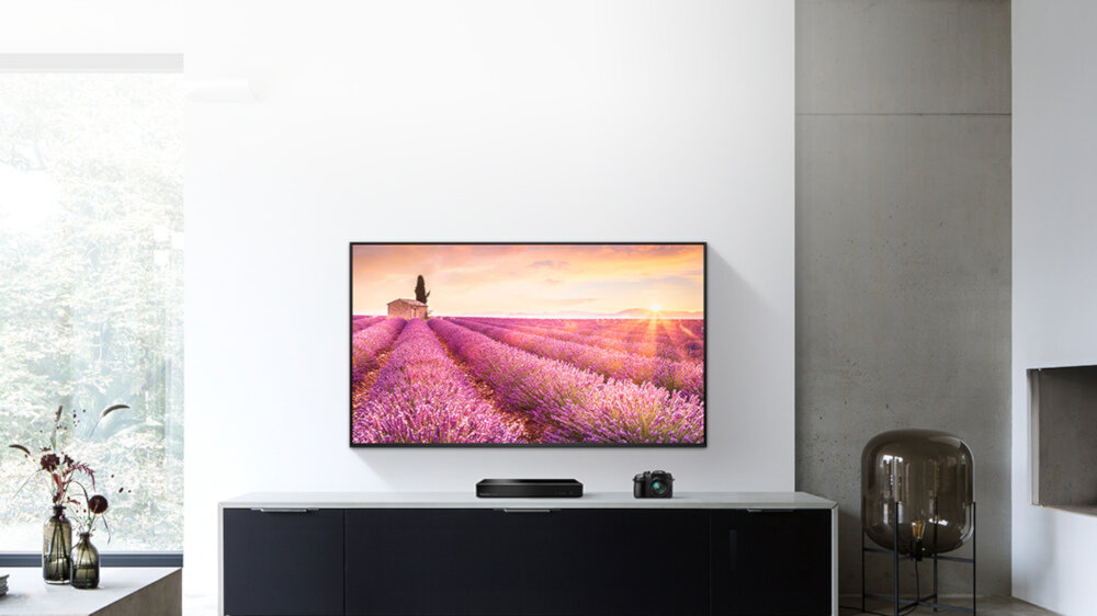 Na ścianie wisi telewizor wyświetlający obraz pola lawendy o zachodzie słońca. Pod telewizorem znajduje się szafka z odtwarzaczem i aparatem fotograficznym.