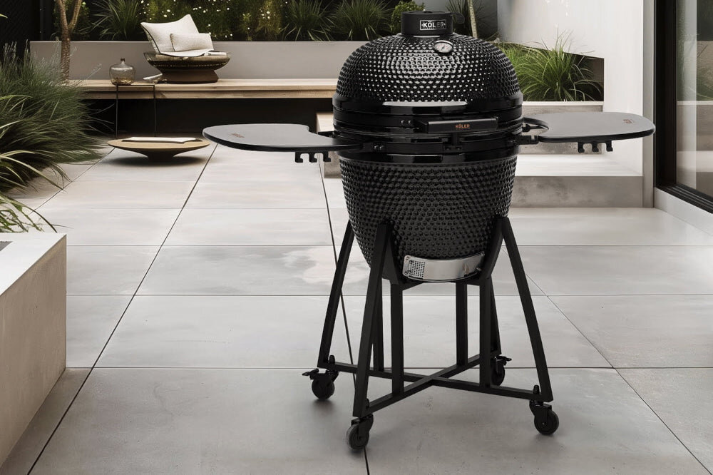 Na tarasie stoi czarny, ceramiczny grill węglowy z okrągłą pokrywą, bocznymi półkami i kółkami. Na pokrywie widoczne jest logo \'KOLER\'.
