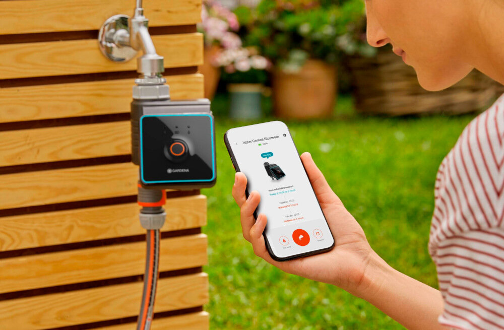 Osoba trzyma smartfon z aplikacją pokazującą urządzenie GARDENA, obok zamontowanego na drewnianej ścianie zaworu z logo GARDENA. W tle widoczny jest ogród z zieloną trawą i roślinami.