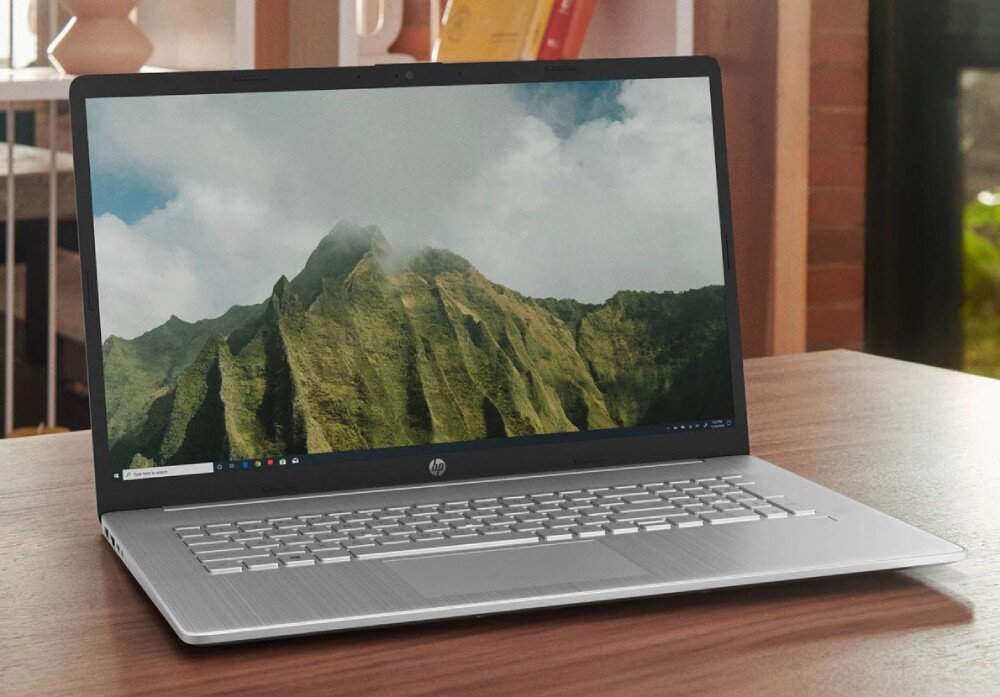Na drewnianym stole stoi otwarty laptop z widocznym logo HP na obudowie. Na ekranie wyświetlany jest krajobraz górski z chmurami.
