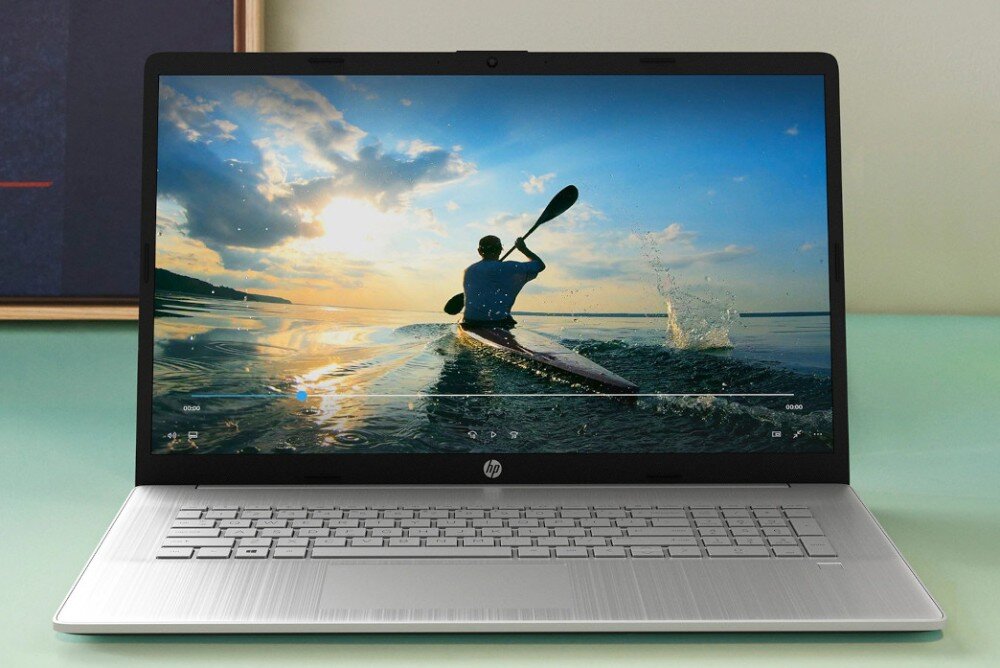 Laptop z wyświetlaczem pokazującym osobę w kajaku na wodzie o zachodzie słońca. Na obudowie widoczne jest logo HP.