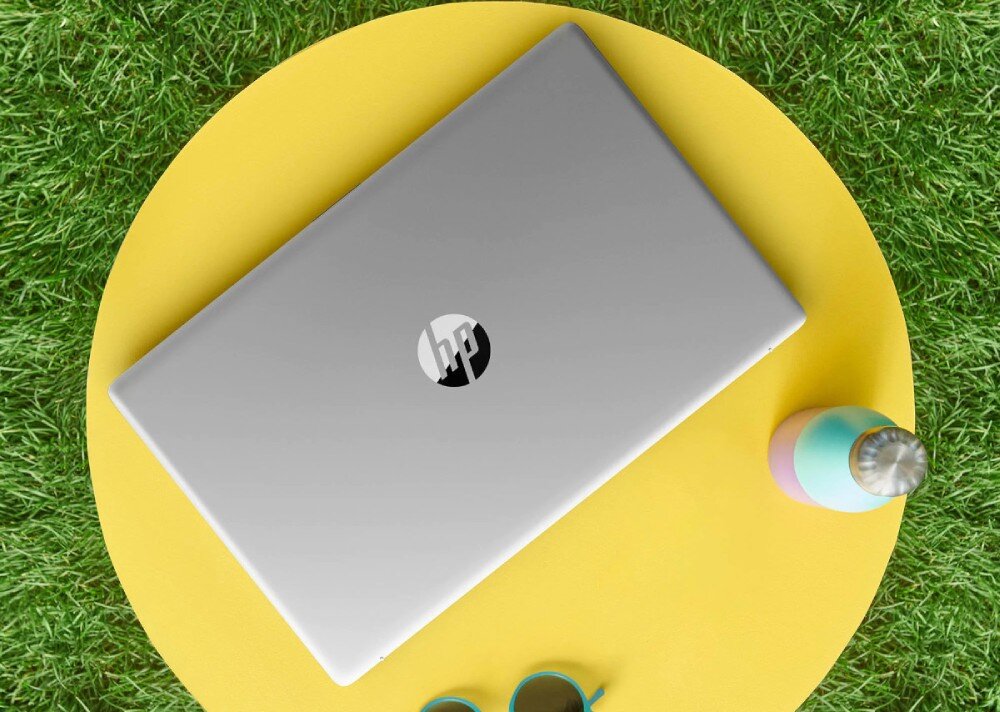 Srebrny laptop z logo \'hp\' na pokrywie leży na żółtym okrągłym tle, otoczony zieloną trawą. Obok znajduje się kolorowa butelka i okulary przeciwsłoneczne.