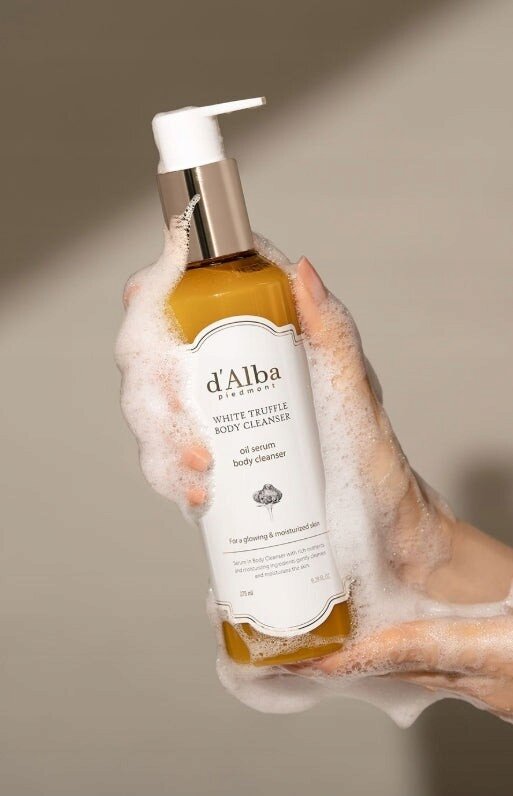 Ręka pokryta pianą trzyma butelkę z pompką, na której etykiecie widnieje napis: \'d\'Alba piedmont WHITE TRUFFLE BODY CLEANSER oil serum body cleanser\'.