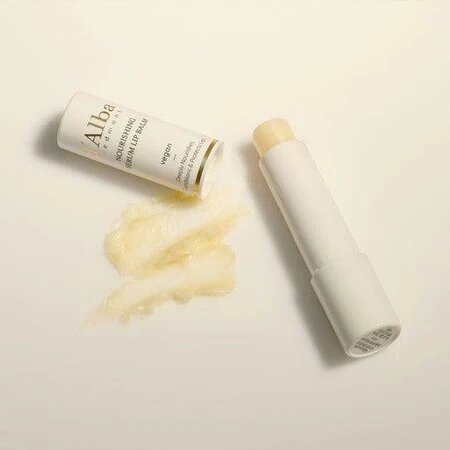 Na obrazie widoczny jest biały balsam do ust w sztyfcie oraz jego opakowanie z napisem \'Alba White Truffle Nourishing Serum Lip Balm\'. Obok sztyftu znajduje się niewielka ilość rozsmarowanego balsamu.