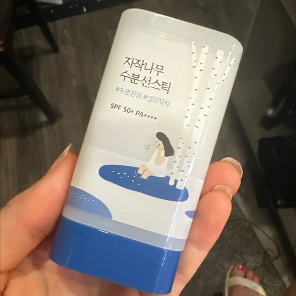 Opakowanie sztyftu z ilustracją kobiety siedzącej przy wodzie i drzewami brzozy. Widoczny tekst: \'SPF 50+ PA++++\'.