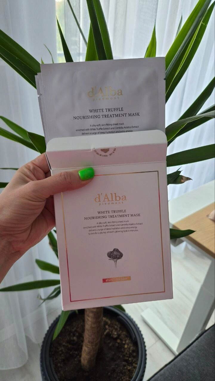 Osoba trzyma opakowanie i saszetkę produktu \'d\'Alba White Truffle Nourishing Treatment Mask\'. W tle widoczna jest roślina doniczkowa i zasłona.