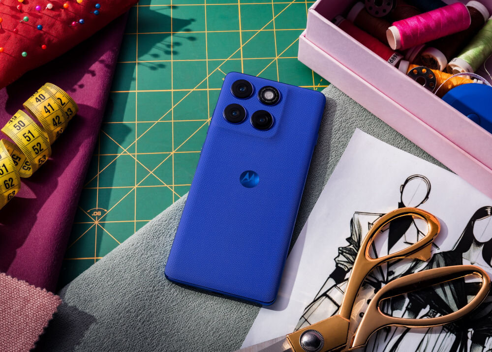 Na zielonej macie do cięcia leży niebieski smartfon z trzema aparatami, obok znajdują się nożyczki, miara krawiecka i pudełko z nićmi. Na telefonie widoczne jest logo w kształcie litery M.