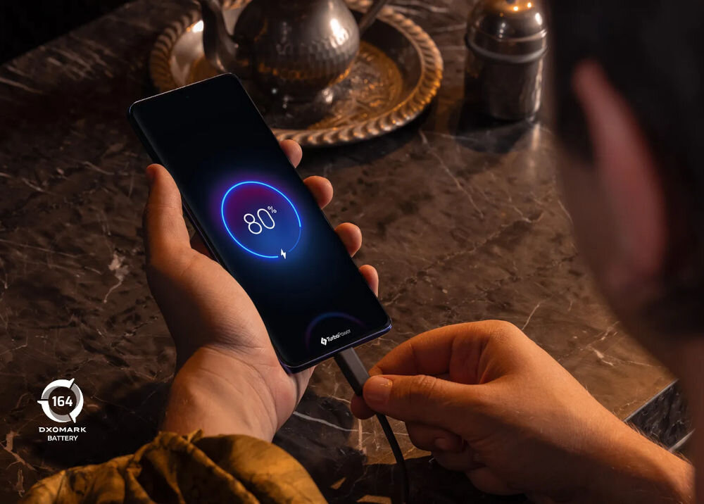 Osoba trzyma smartfon z wyświetlaczem pokazującym 80% naładowania baterii. W lewym dolnym rogu widoczny jest napis \'164 DXOMARK BATTERY\'.