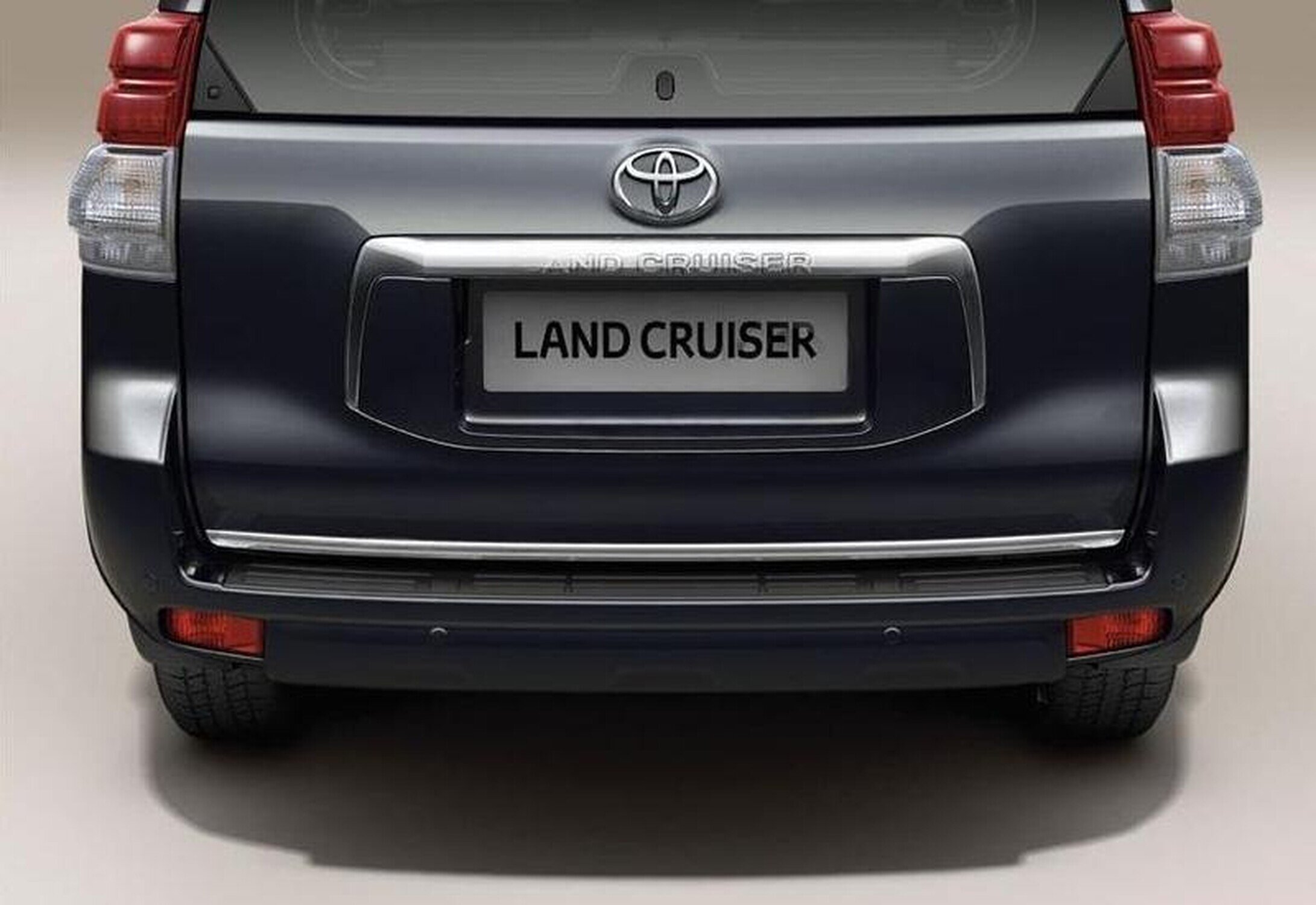 Zdjęcie przedstawia tył samochodu z widocznym logo Toyota i napisem \'LAND CRUISER\' na tablicy rejestracyjnej. Samochód ma ciemny kolor i chromowane elementy na zderzaku.
