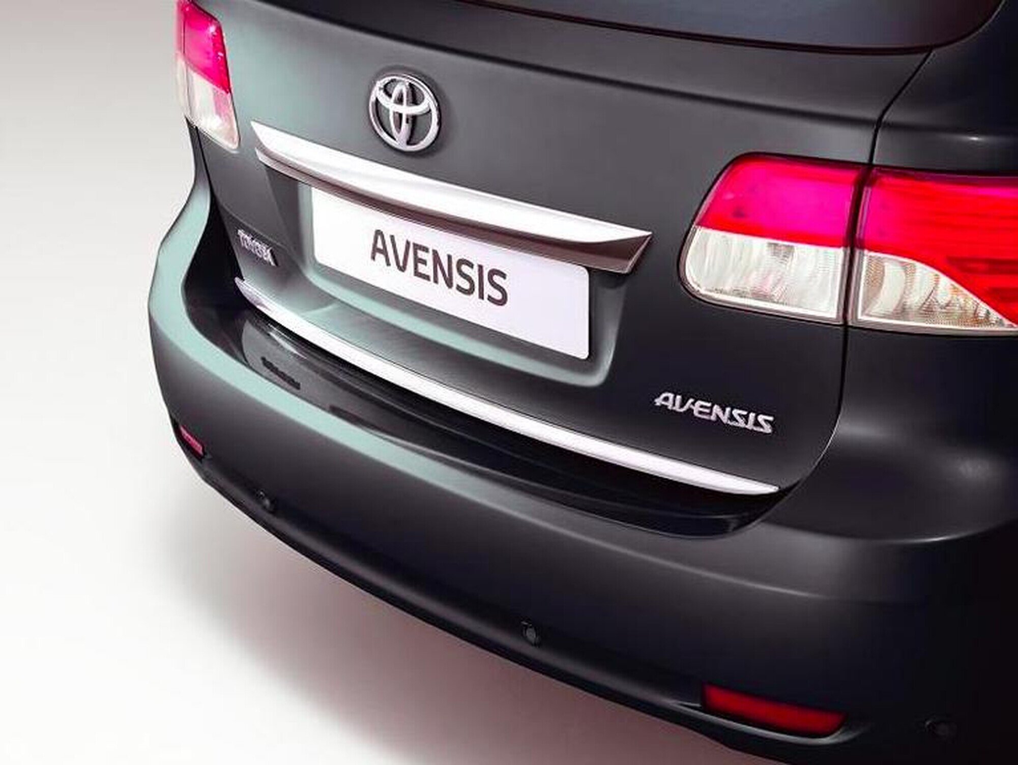Zdjęcie przedstawia tylną część samochodu z widocznym logo Toyota oraz napisem \'AVENSIS\' na tablicy rejestracyjnej. Samochód ma ciemny kolor, a na zderzaku znajduje się srebrna listwa ochronna.