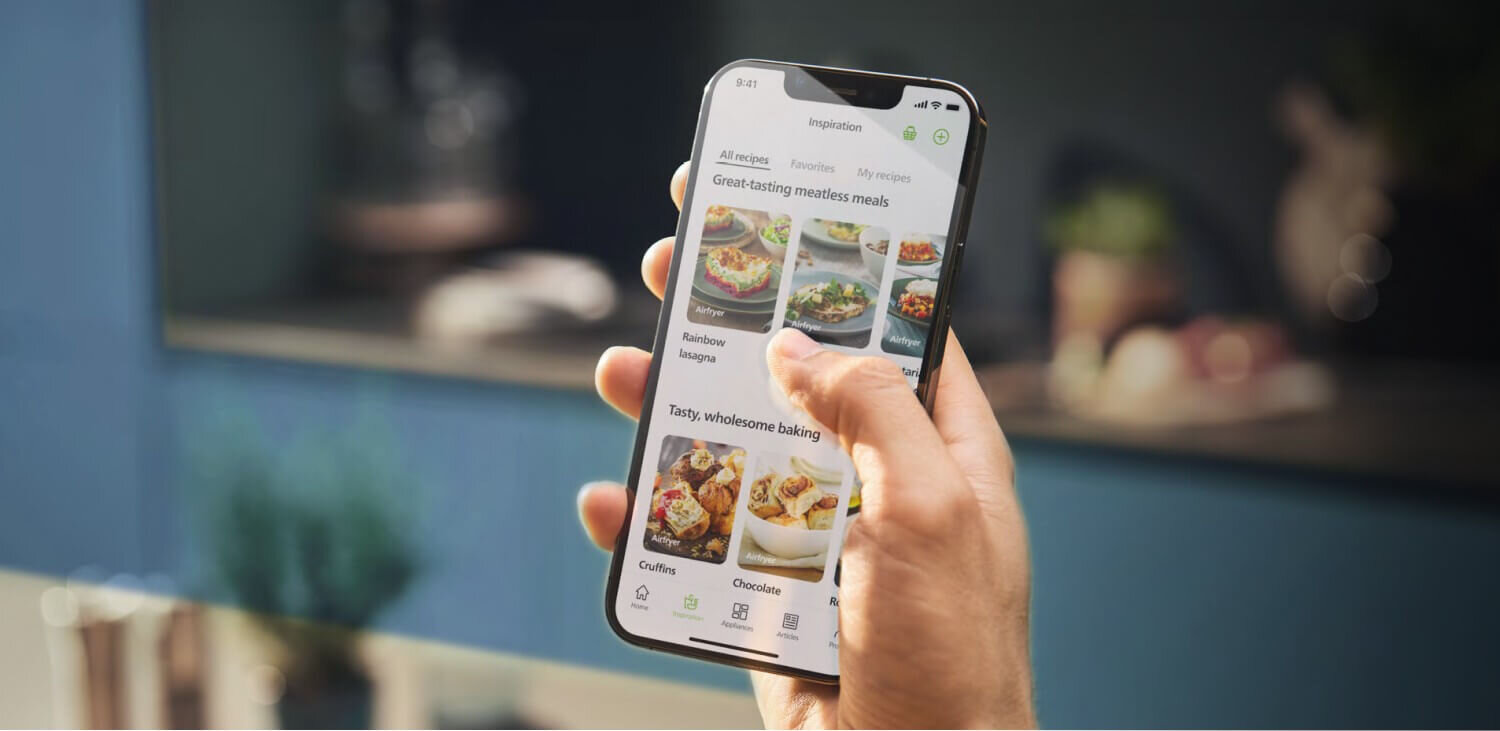 Osoba trzyma smartfon, na ekranie którego widoczna jest aplikacja z przepisami kulinarnymi, zawierająca zdjęcia potraw i teksty takie jak \'Great-tasting meatless meals\'.