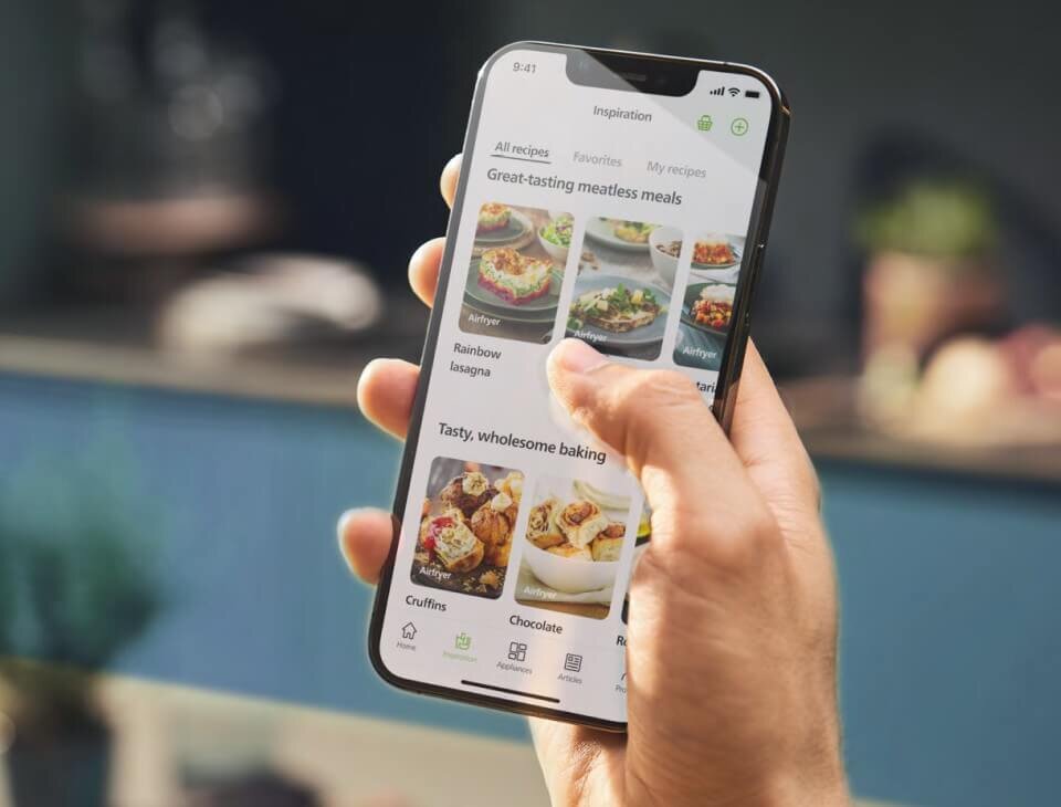 Osoba trzyma smartfon, na którym wyświetlana jest aplikacja z przepisami kulinarnymi. Na ekranie widoczne są zdjęcia potraw oraz teksty takie jak \'Great-tasting meatless meals\' i \'Tasty, wholesome baking\'.