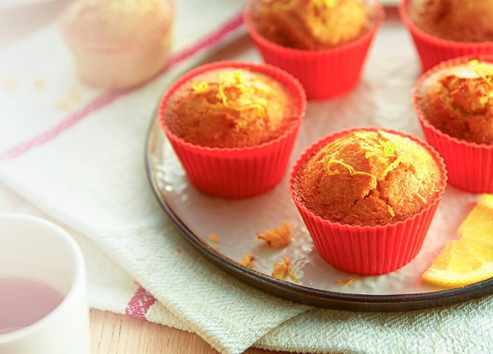 Na talerzu znajdują się muffiny w czerwonych silikonowych foremkach, ozdobione skórką cytrynową. Obok talerza widoczna jest filiżanka z napojem.
