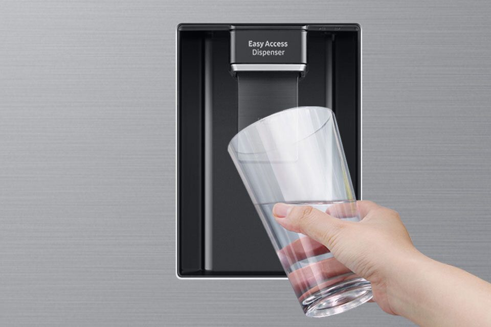 Łatwy w obsłudze dystrybutor wody - Samsung RB38C635ES9 Osoba trzyma szklankę pod podajnikiem wody z napisem \'Easy Access Dispenser\' na tle szczotkowanej stali nierdzewnej.