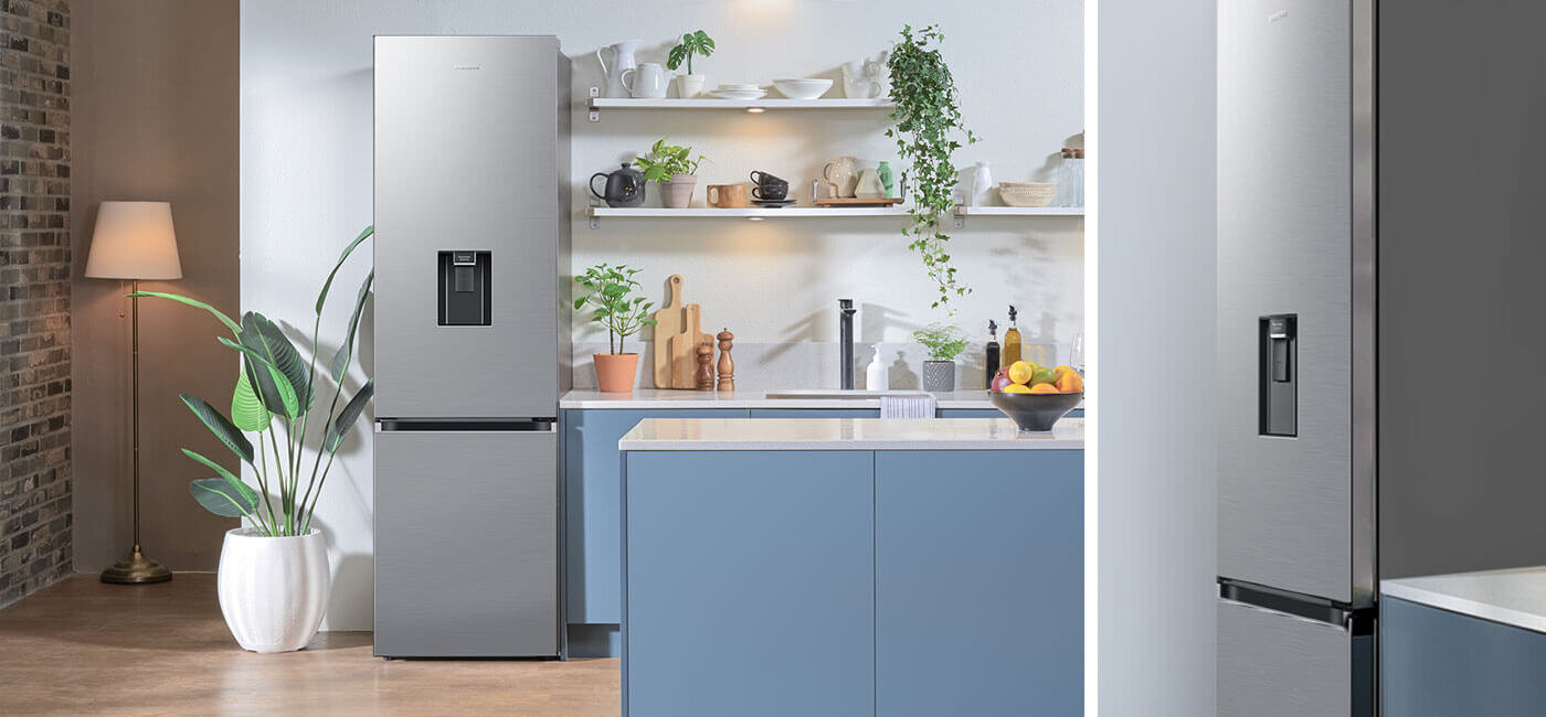 Built-In dla lodówki Samsung RB38C635ES9 W kuchni znajduje się lodówka w kolorze szczotkowanej stali nierdzewnej z podajnikiem wody na drzwiach. Wnętrze kuchni jest nowoczesne, z niebieskimi szafkami i roślinami na półkach.