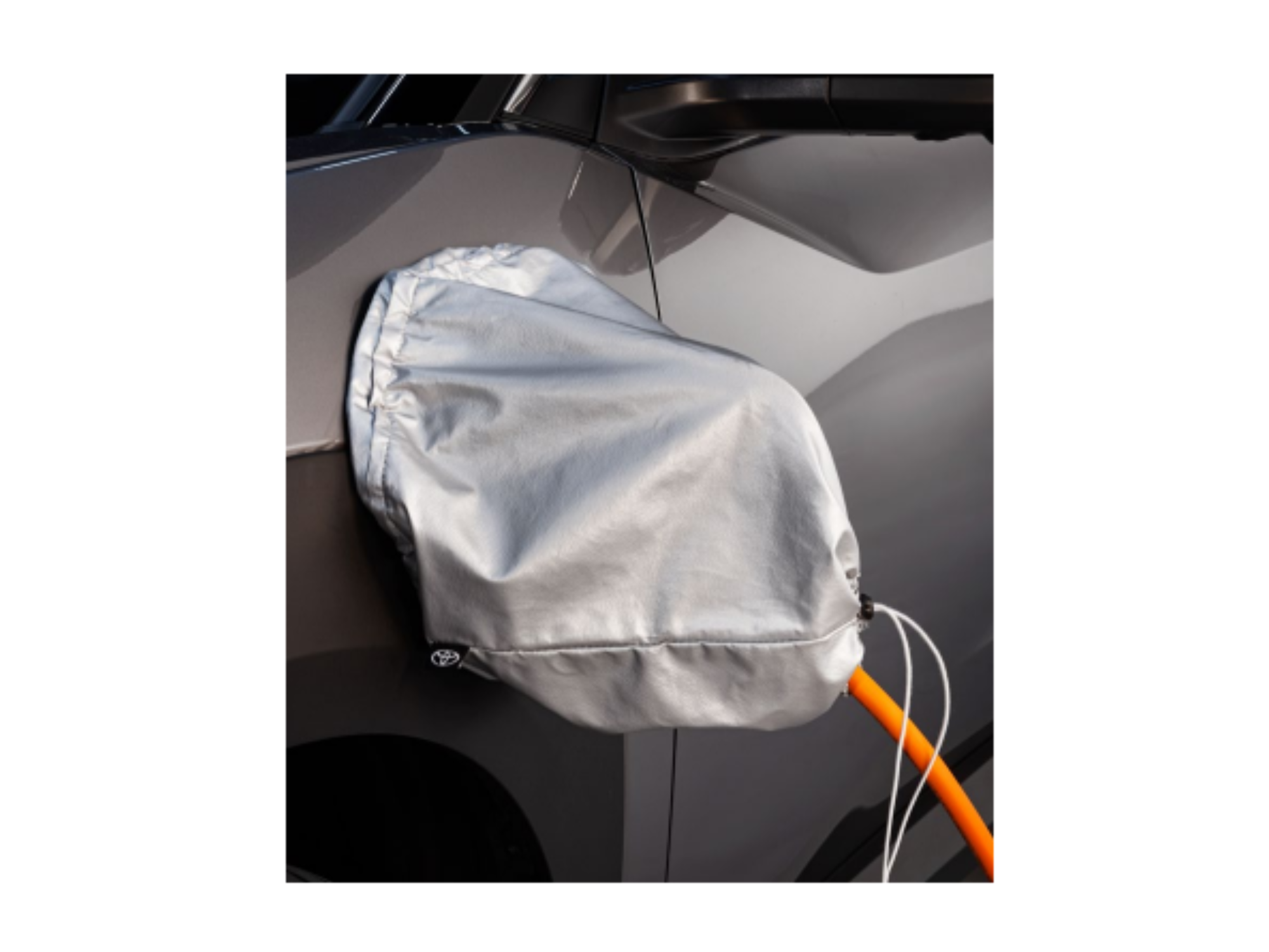 Na obrazie widoczny jest szary pokrowiec zabezpieczający gniazdo ładowania samochodu, z którego wystaje pomarańczowy kabel. Na pokrowcu znajduje się logo Toyota.