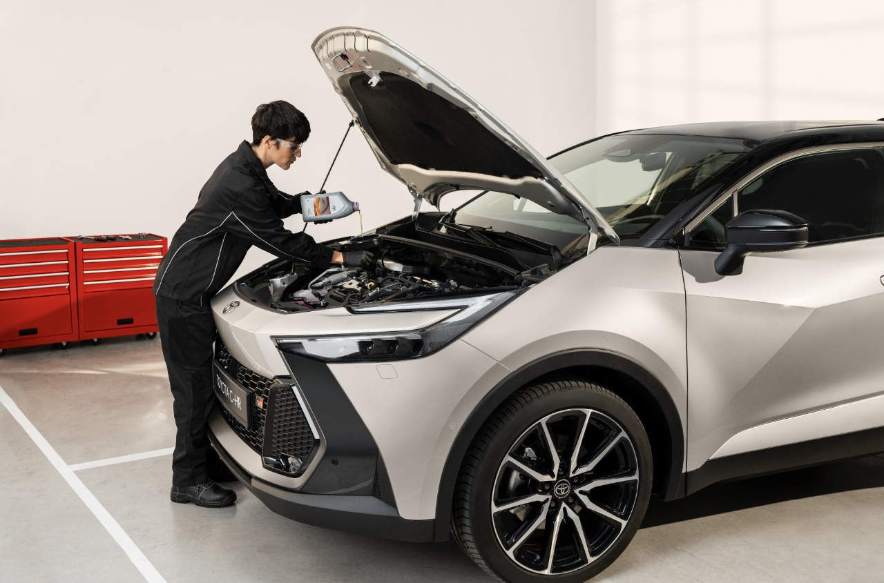 Osoba w stroju roboczym sprawdza silnik samochodu z otwartą maską. W tle widoczne są czerwone szafki narzędziowe.