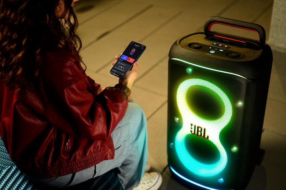 Osoba trzymająca smartfon siedzi obok głośnika z podświetleniem LED w kształcie ósemki i logo JBL. Głośnik ma uchwyt i panel sterowania na górze.