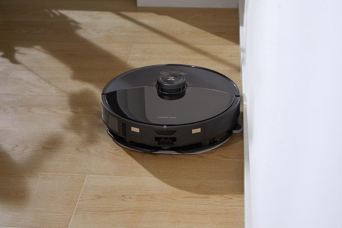 Czarny robot sprzątający znajduje się na drewnianej podłodze przy ścianie. Na urządzeniu widoczne jest logo \'roborock\'.
