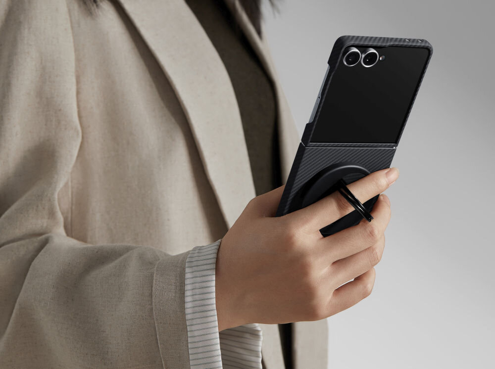 Osoba trzyma telefon w czarno-szarym etui z widocznym okrągłym uchwytem. Telefon ma dwa obiektywy aparatu na tylnej obudowie.