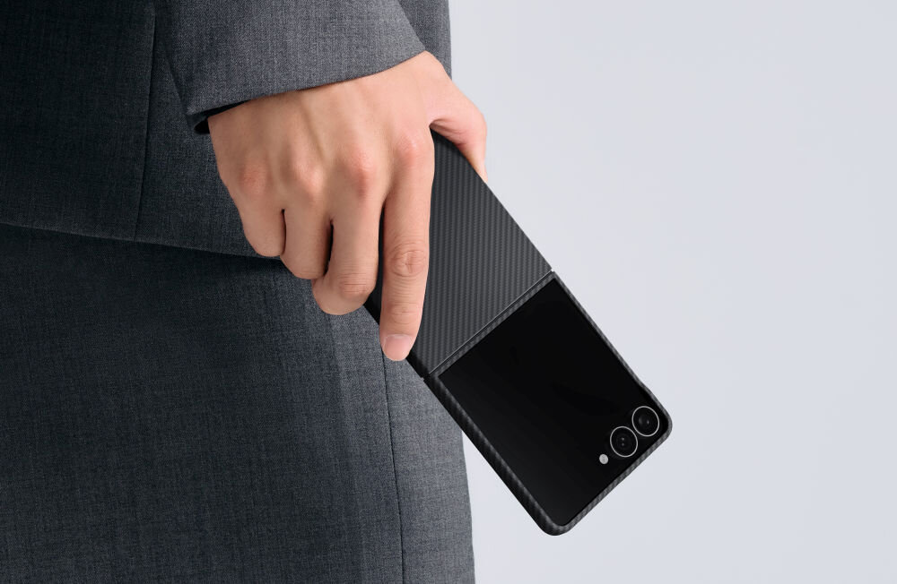 Osoba trzyma w ręce telefon w czarno-szarym etui z widocznymi dwoma obiektywami aparatu. Tło jest jednolite i jasne.