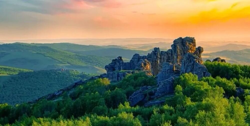 Na obrazie widoczny jest malowniczy krajobraz z formacjami skalnymi otoczonymi zielonymi drzewami, a w tle rozciągają się wzgórza pod pomarańczowym niebem o zachodzie słońca.