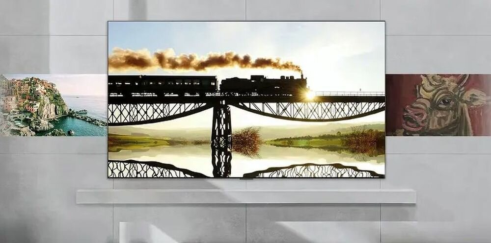 Telewizor wyświetla obraz pociągu jadącego po moście z odbiciem w wodzie poniżej. Po bokach telewizora widoczne są fragmenty obrazów przedstawiających krajobraz i głowę krowy.