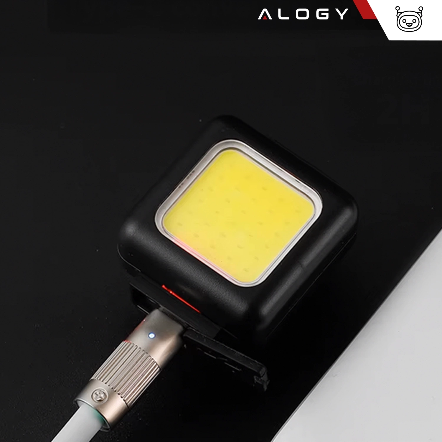Kwadratowa lampka rowerowa z żółtym panelem świetlnym i czarną obudową, podłączona do kabla USB. W górnym rogu widoczne jest logo \'ALOGY\'.