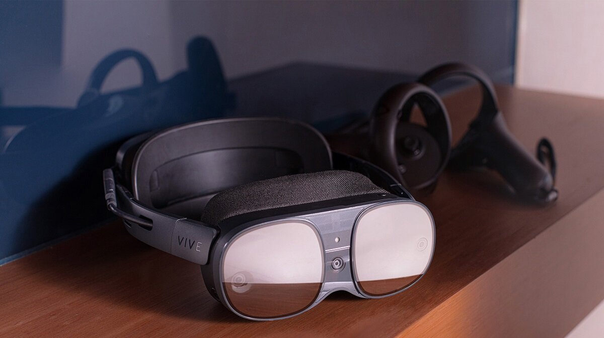 Na drewnianej powierzchni leżą gogle VR z widocznym napisem \'VIVE\' oraz kontroler. W tle znajduje się ciemna powierzchnia, prawdopodobnie ekran.