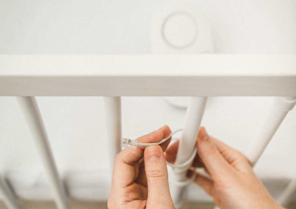 Osoba trzyma kabel z wtyczką, który jest przeciągany przez szczebelki białego łóżeczka. W tle widoczny jest biały, okrągły element zamontowany na łóżeczku.