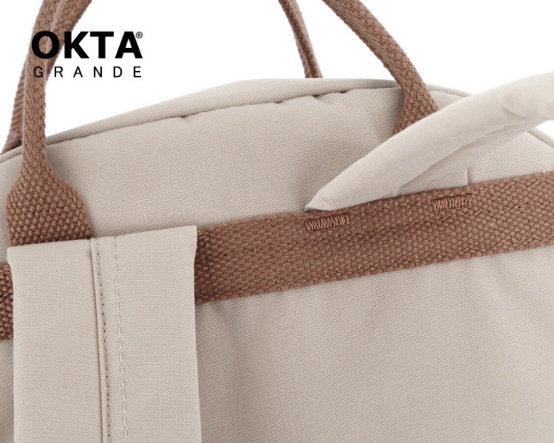 Fragment plecaka w kolorze beżowym z brązowymi uchwytami. W lewym górnym rogu widoczne jest logo \'OKTA GRANDE\'.