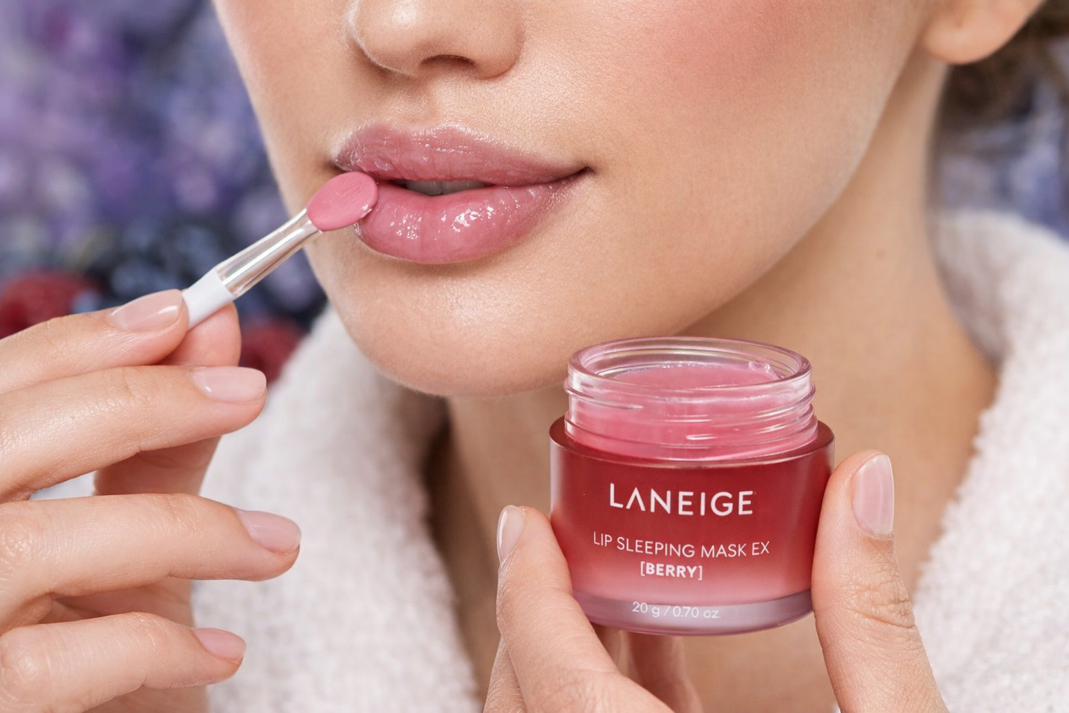 Osoba aplikuje różowy produkt na usta za pomocą małego pędzelka. Widoczny jest słoiczek z napisem \'LANEIGE LIP SLEEPING MASK EX [BERRY] 20g/0.70oz\'.