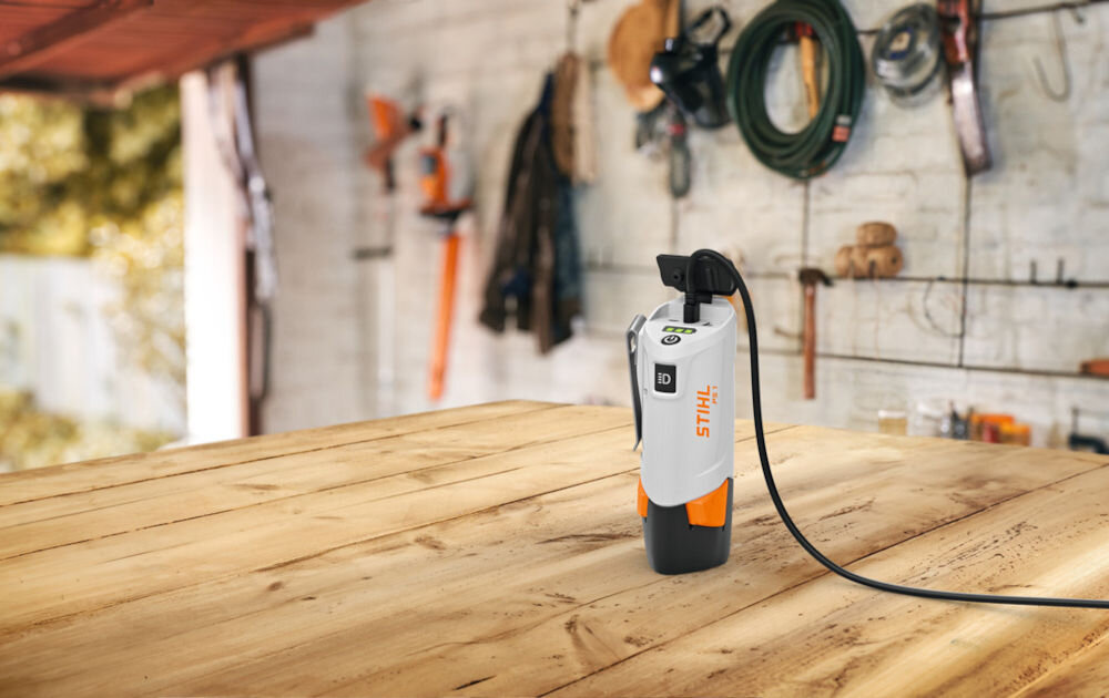 Na drewnianym stole w warsztacie znajduje się powerbank z logo STIHL, podłączony kablem. W tle widoczne są narzędzia i sprzęt ogrodniczy zawieszone na ścianie.