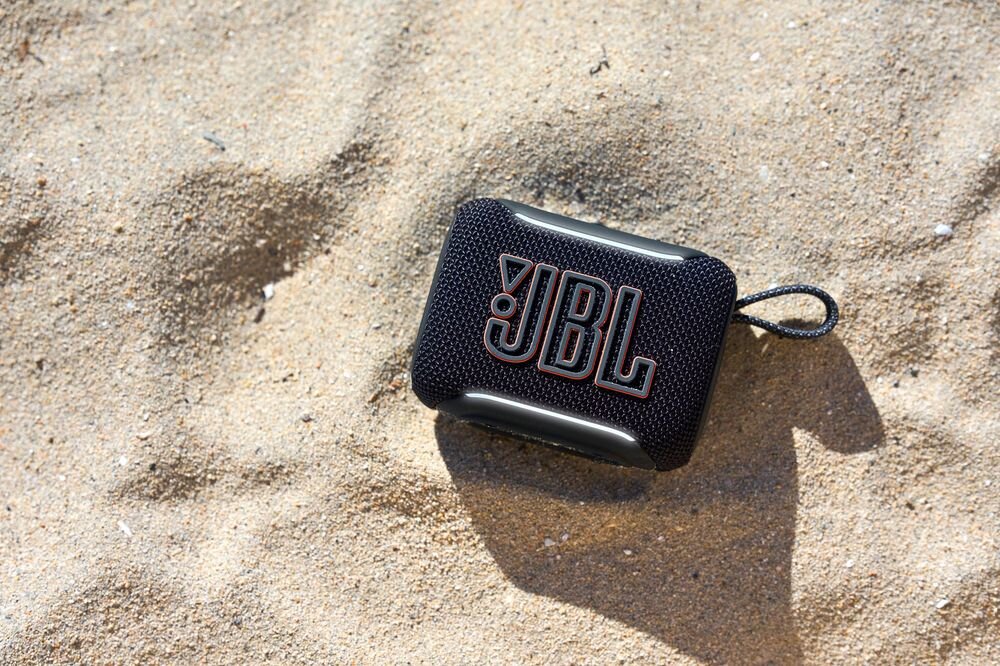 Czarny głośnik mobilny z logo \'JBL\' leży na piasku. Głośnik ma teksturowaną powierzchnię i pętlę do przenoszenia.