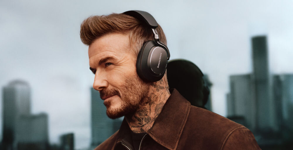 Osoba nosi czarne słuchawki nauszne z widocznym logo \'Bowers & Wilkins\'. W tle znajduje się rozmyty widok miasta.