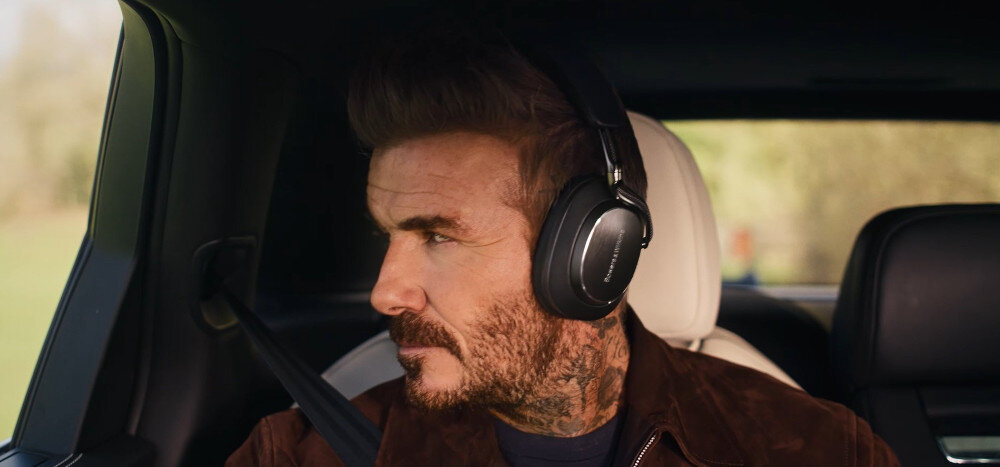 Osoba siedzi w samochodzie, nosząc czarne słuchawki nauszne z widocznym logo \'Bowers & Wilkins\'.