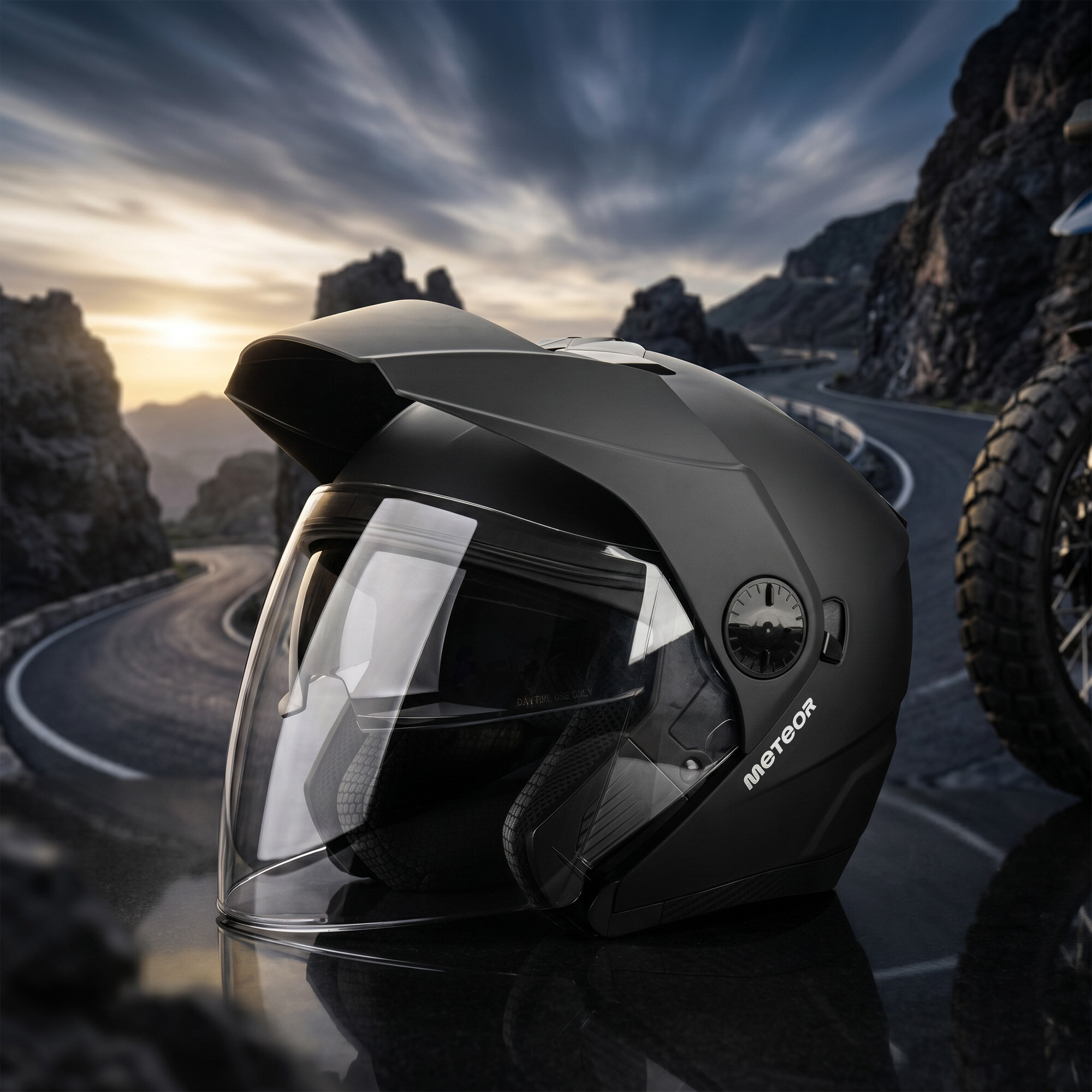 Czarny kask motocyklowy z przezroczystą szybką, na tle krętej górskiej drogi. Na kasku widoczne jest logo \'METEOR\'.